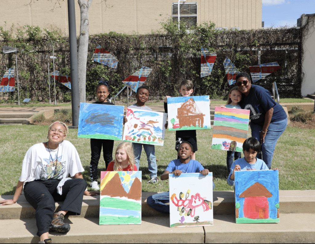 A group of children proudly holding their art along with 2 adults outside on a sunny day